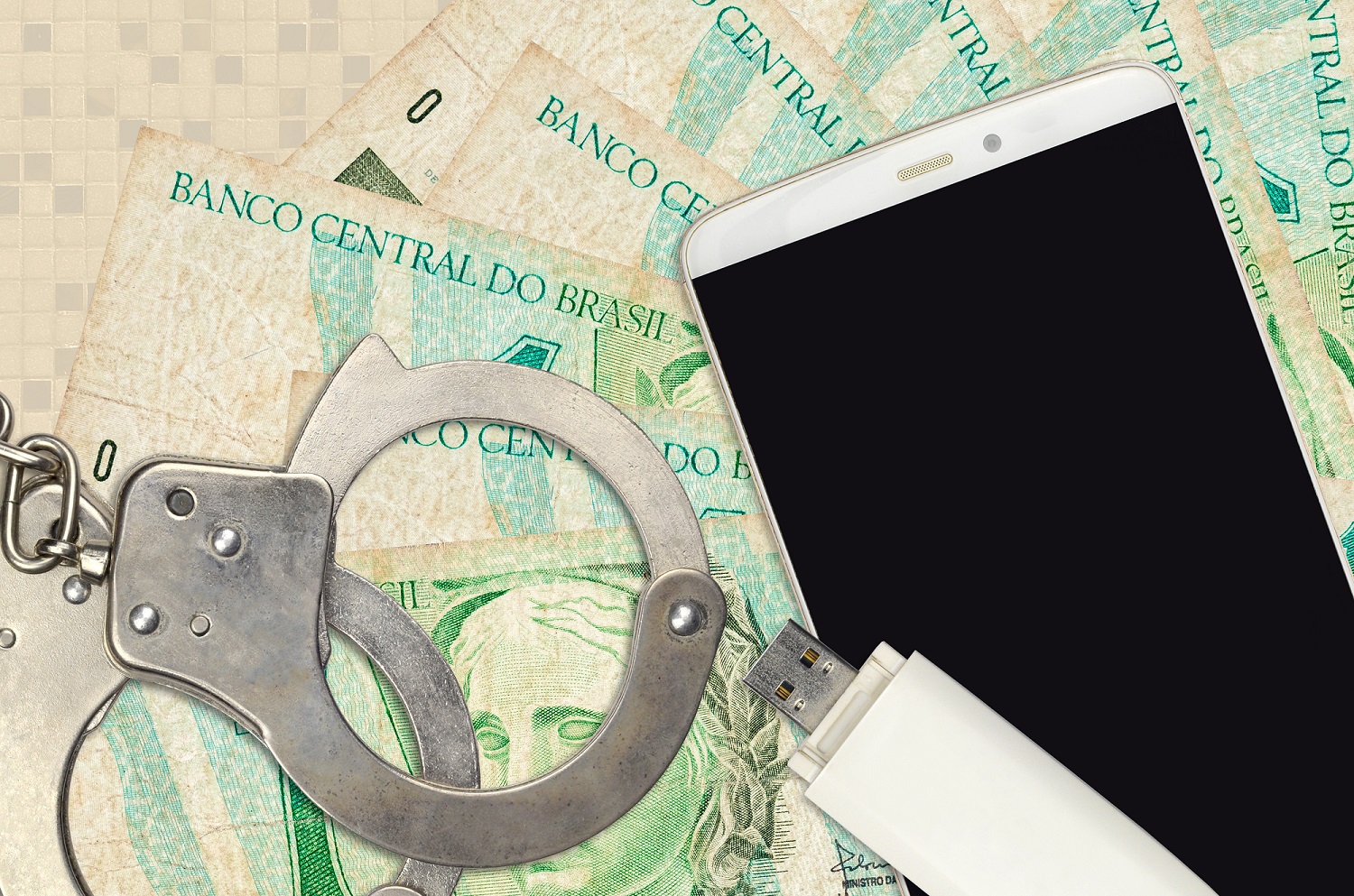 На куче бразильских банкнот лежат USB-накопитель и смартфон вместе с парой наручников, а также фотографии предполагаемых мошенников.