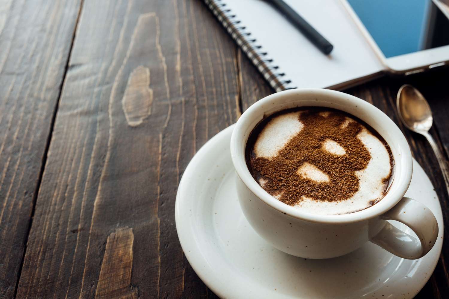 Чашка кофе на деревянном столике рядом с блокнотом и мобильным телефоном, украшенная корицей в виде логотипа Bitcoin.
