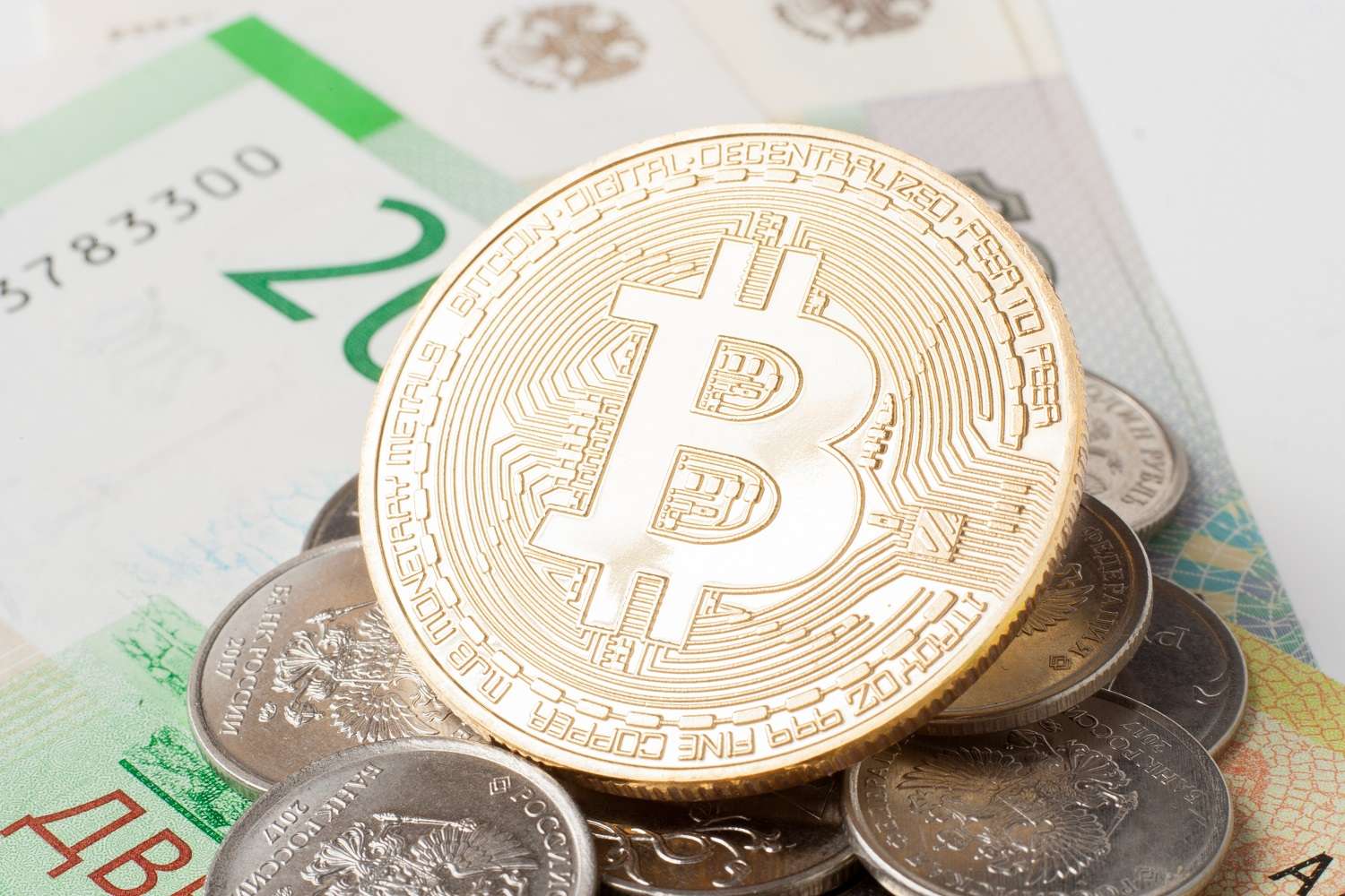 Металлический токен, символизирующий Bitcoin, лежит на вершине стопки российских монет и банкнот.