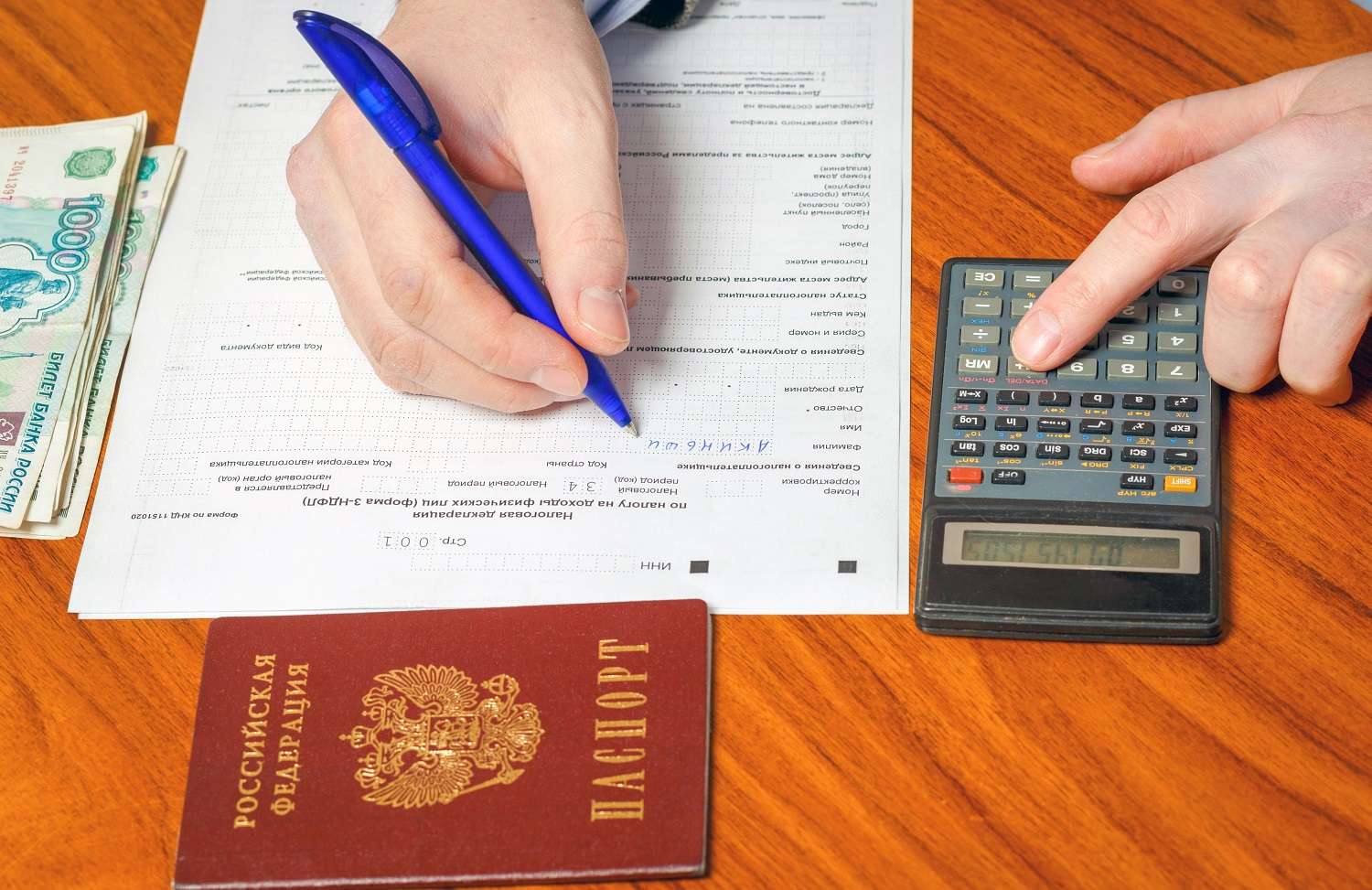 A person fills in a Russian tax return, holding a pen. Next to the form is a small pile of Russian banknotes and a calculator. The person presses a key on the calculator.